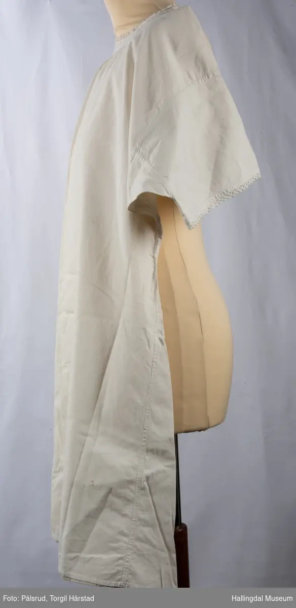 Hvit nattkjole eller serk i bomull med heklet kant rundt ermer og hals. Et hull er bøtt med en firkantet, hvit stoffbit midt framme nederst.
