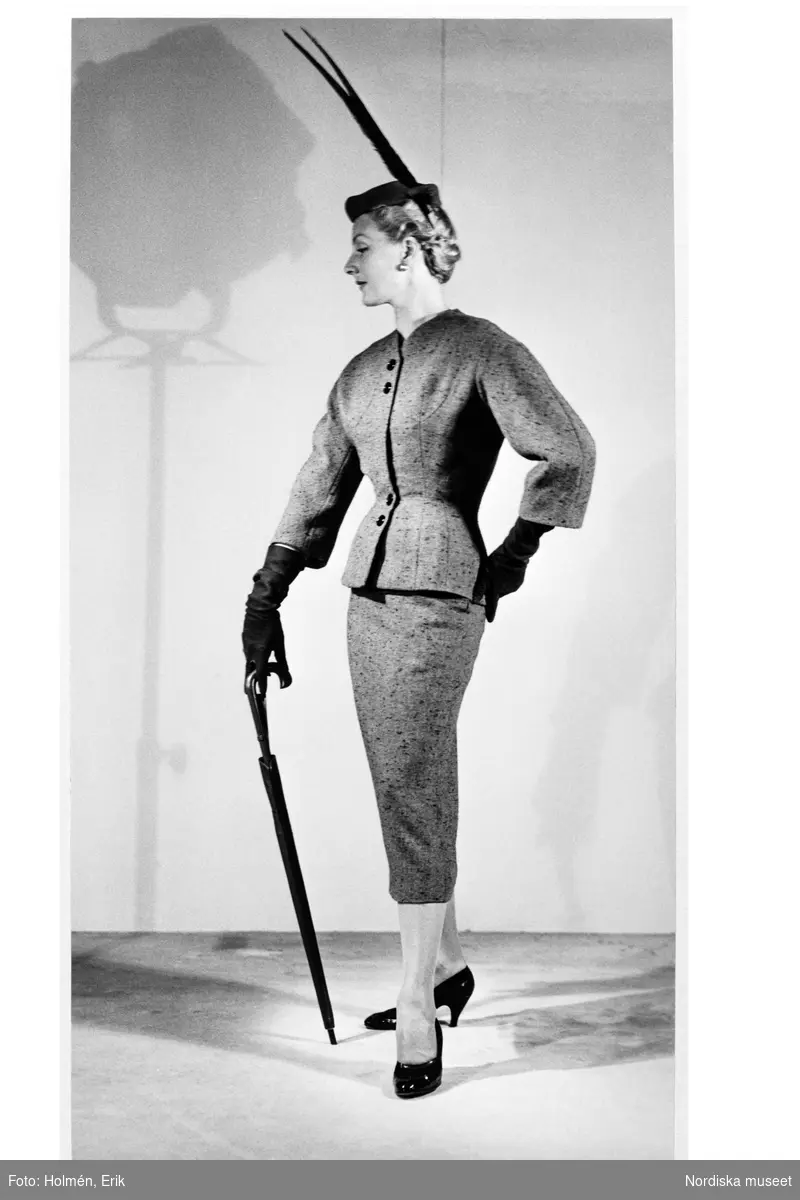 Mannekängen Margareta Berglund gift Nordlund (1946) visar dräkt i provrummet vid NK:s Franska damskrädderi. Figursydd jacka, stusslång med vidd vid byst och den trekvartslånga ärmen. Vadlång pennkjol. Hatt med långa fjädrar, handskar och lackpumps. Paraply i den ena handen.