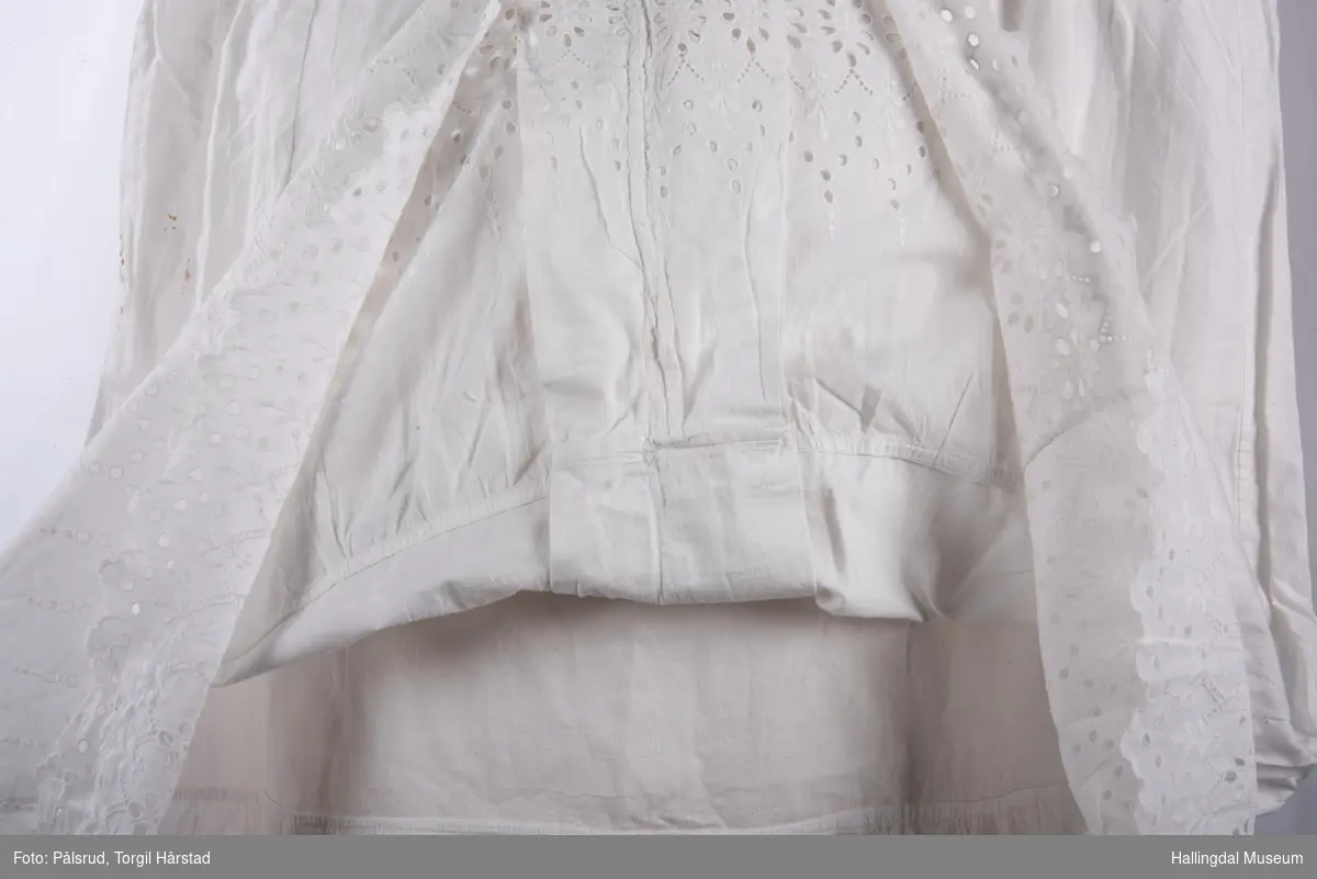 Hvit underkjole i bomull med bred heklet kant øverst på brystet rundt hals og ermer. To heklet stropper. Bred heklet kant nederst på kjolen. Fasongsydd.