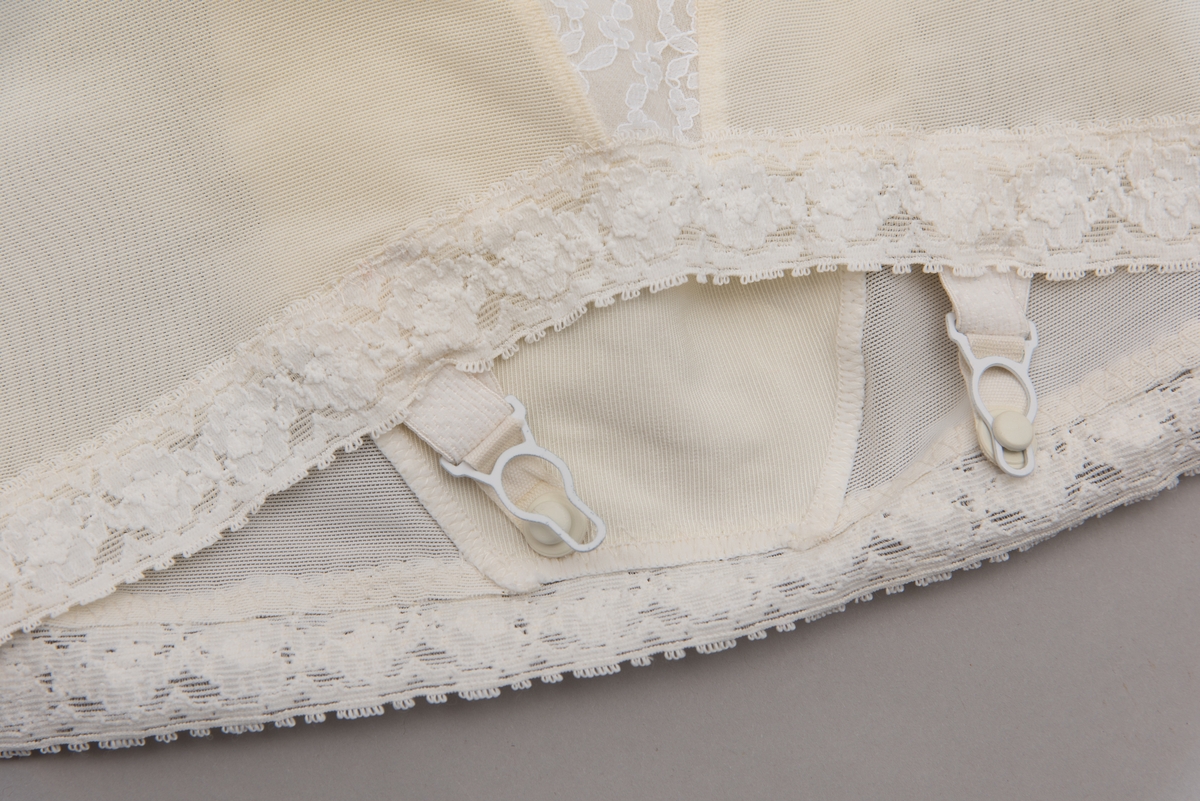 Hvit hofteholder med stropper og blondekant nederst og foran på midtstykke. Pyntet med sløyfe og kulørt blomst i tekstil foran på linning.