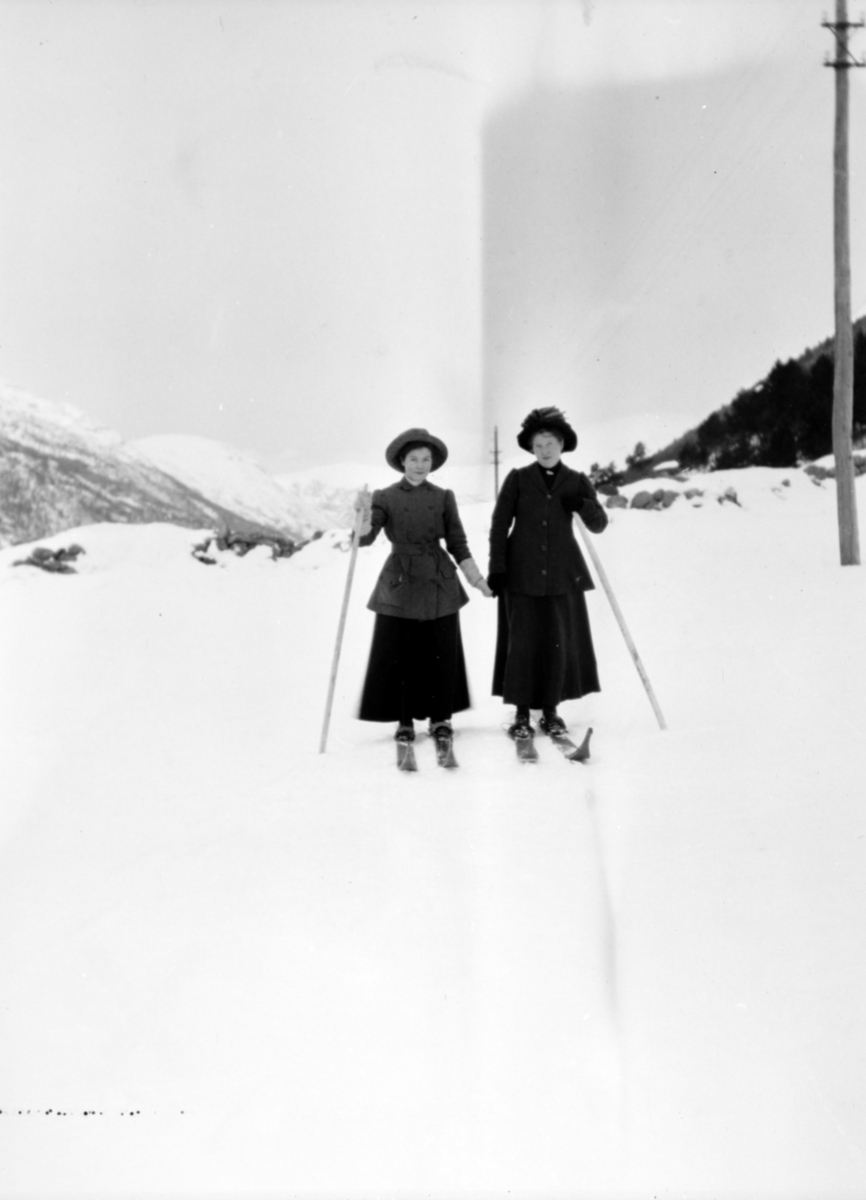 To damer som går på ski med en stav. Begge er kledd i lange stakker, lange jakker og hatt. Bildet er fra Kyllingjordet på Verma med Sæterhøa og Døntefjellet i bakgrunn