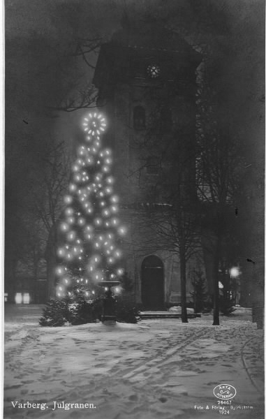 Vykort, "Varberg. Julgranen". Kvällsbild av en tänd julgran i Brunnsparken med kyrktornet i bakgrunden. Marken är täckt av snö. Stämpeln nere till höger försäkrar att kortet är "Äkta fotografi", foto & förlag: Björkström.