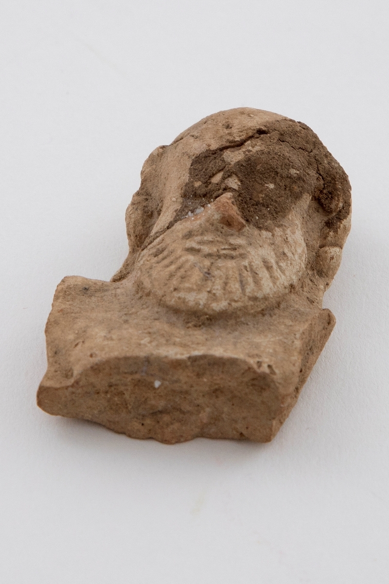 Fragment av figurin i terrakotta, øvre del av kroppen er bevart. Fremstiller en faun med skjegg. Synlige brudd og rester av jord.