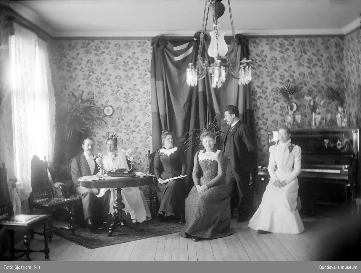 Gruppfoto, familjeporträtt i hemmiljö. Ett brudpar samt ytterligare tre kvinnor och en man.