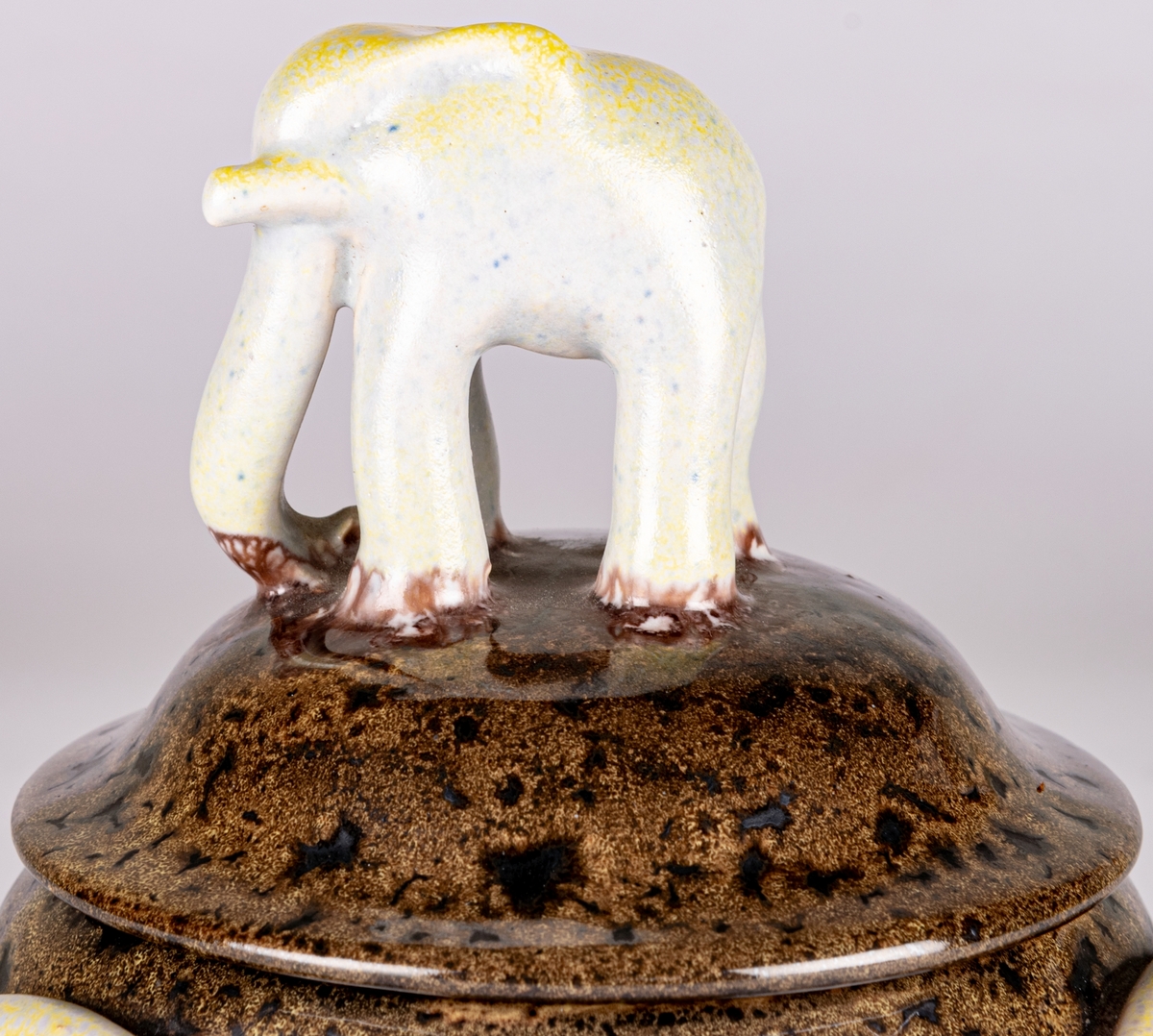 Bonbonjär, konfektskål med lock, formgiven av Allan Ebeling. Brunspräcklig glasyr, tre dekorationsöron på sidorna i en gulskimrande glasyr, locket kröns av en modellerad elefant i en gråblå och gulskimrande glasyr.