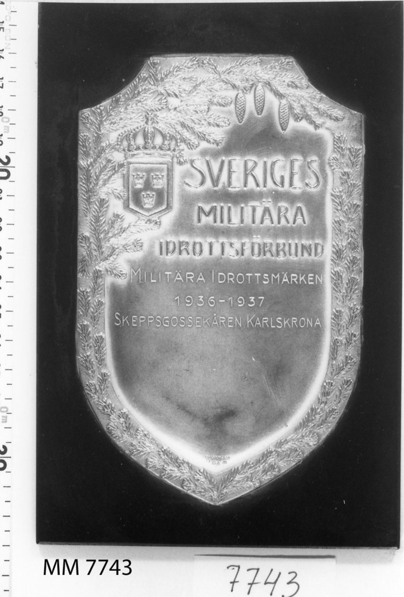 Plakett av silver på platta av trä, svart.
Inskription: Sveriges Militära Idrottsförbund
Militära Idrottsmärke 1936-1937
Skeppsgossekåren Karlskrona.