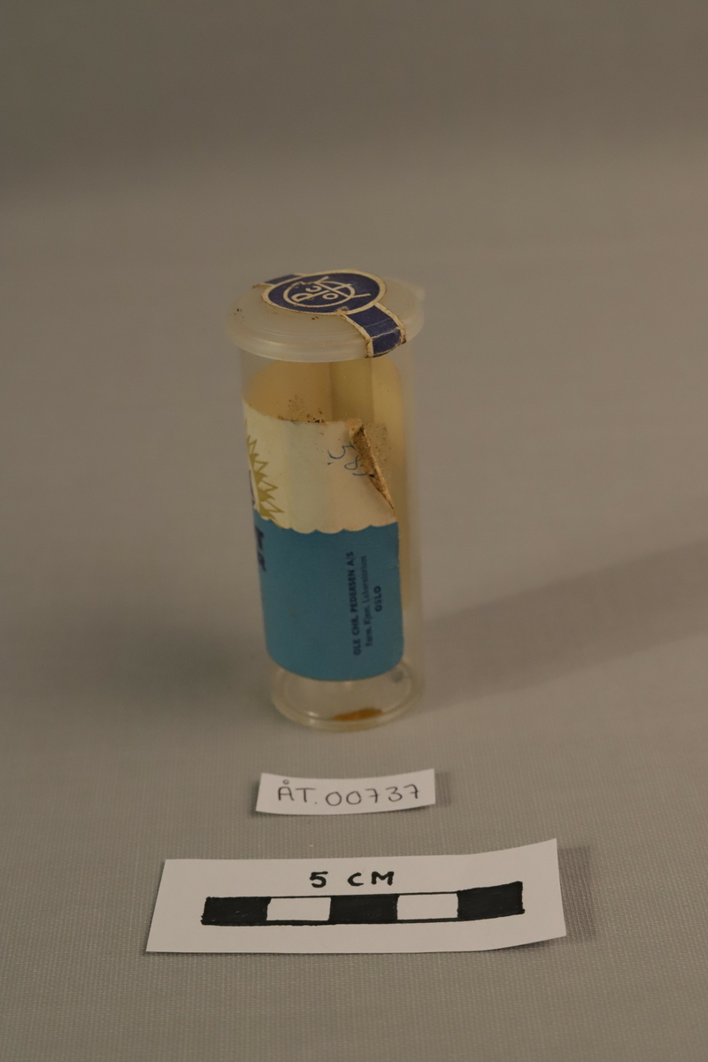 Klar plastikk tube med papir etikett i gull, mørkeblått og lyseblått motiver og skrift.