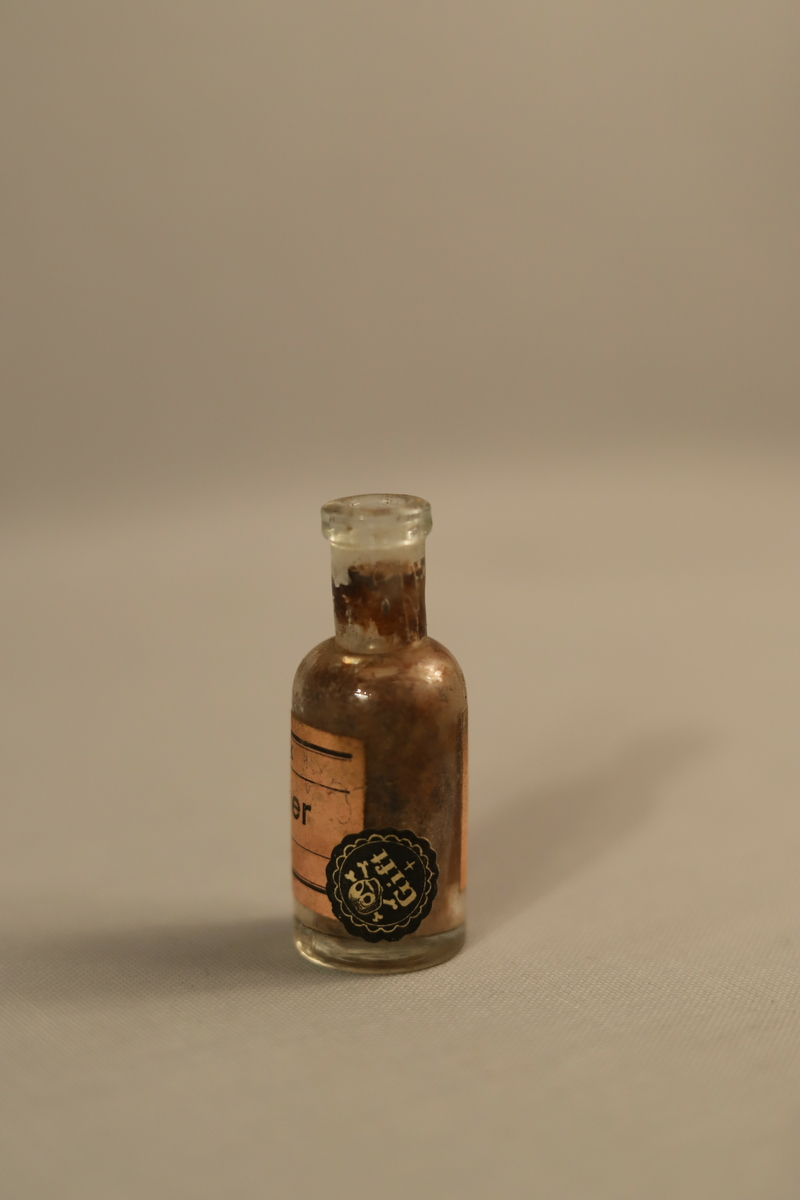 Klar glass flaske og beige etikett med sort skrift. Etiketten er møkkete og slitt. Flasken har mørke resten i seg som får den til å fremstå som sort. 