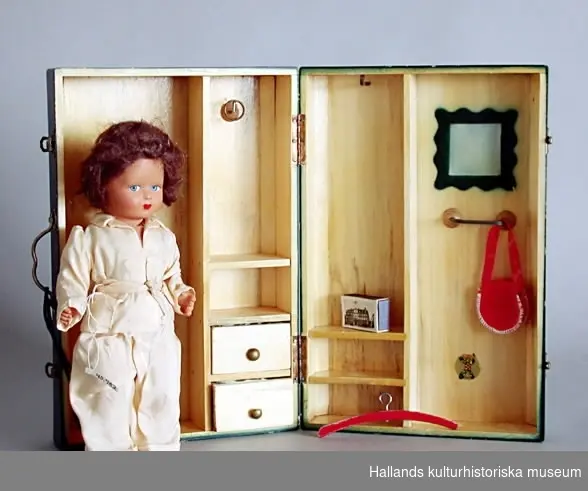 Dockväska av trä, grönmålad. Innehåller två dockor, dockkläder samt tillbehör till dockorna. Uppfälld bildar väskan en dockgarderob.