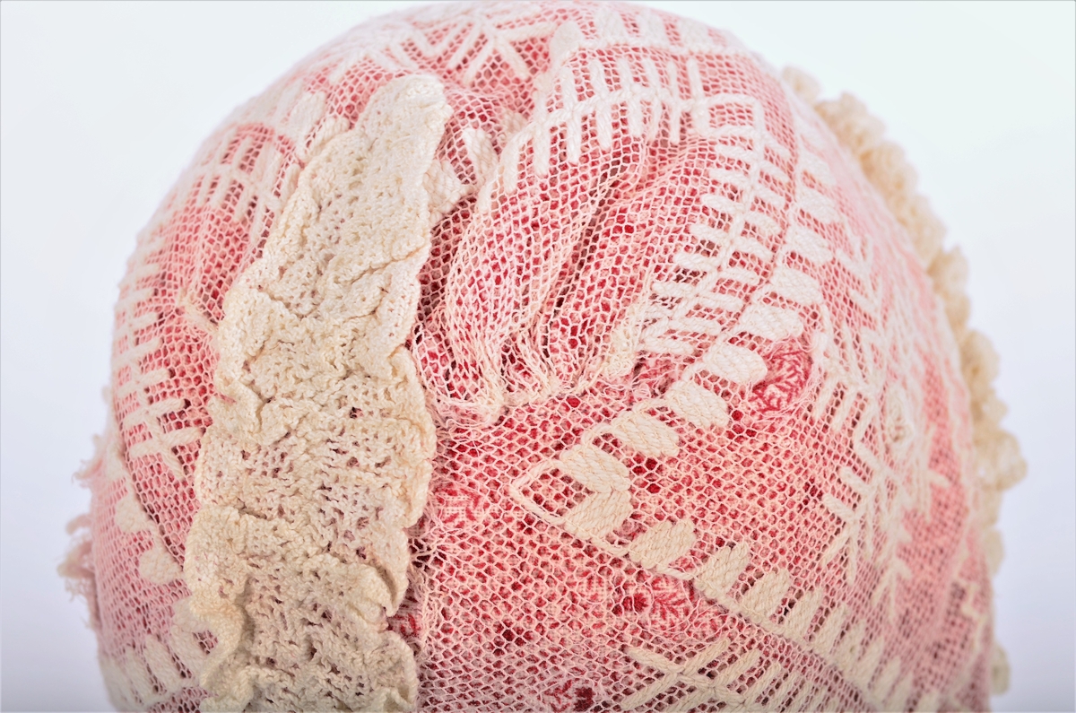 Dåpslue i kvit og raudt mønstra trykt bomullstøy, trekt utanpå med tyllbrodri. Lua er av eit tøystykke med saum midt bak, 9cm høg, I enden av saumen ein samansynka saum (7cm) tverrs over. Påsett blonde over saumen på retta. Dobbel rysjekant langs framkanten og nederst. 12 små rosa silkeband javnt fordelt i rysjekanten framme. To knyteband i silke.
Tyllbroderiet er mest sannsynleg del av ei rullehette.