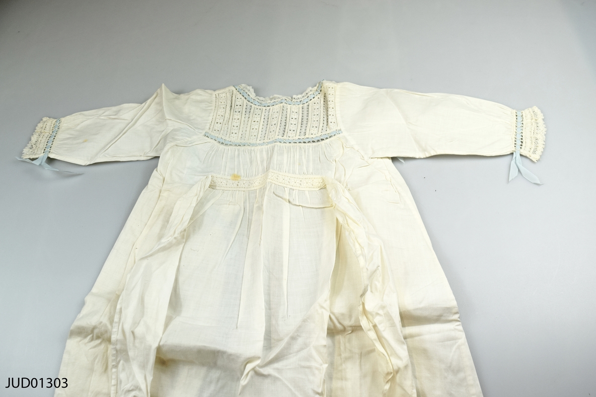 Kläder för barn i blå ask med knytband: vita, broderade klänningar, skjortor, hättor och västar, samt två par skinnhandskar i damstorlek.