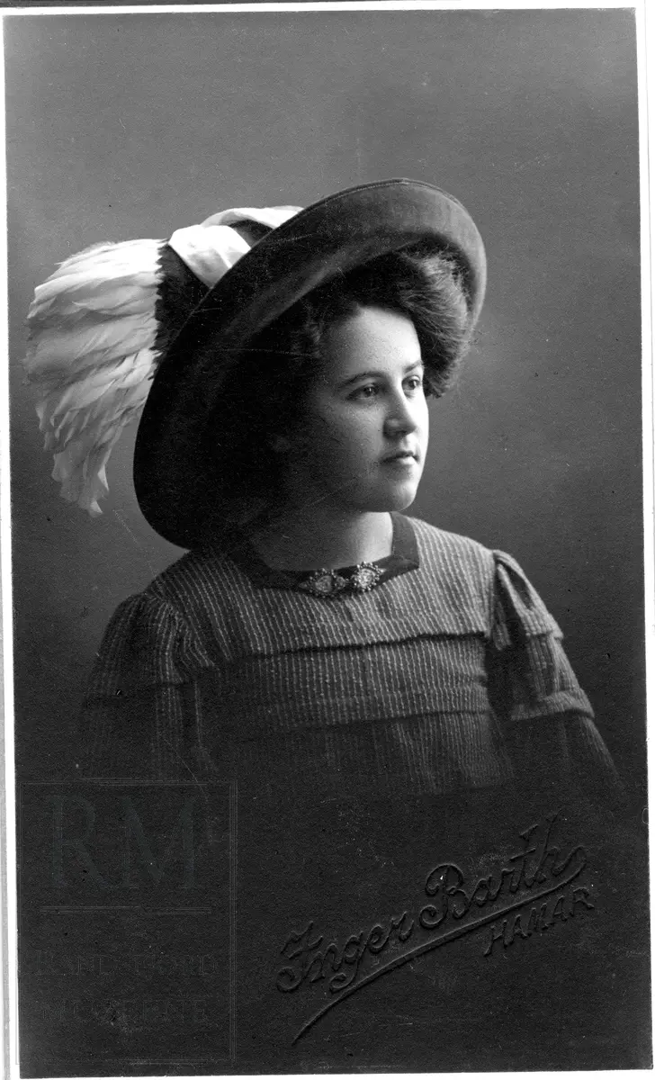 Brystbilde av en ung kvinne med stor hatt med fjær. I halsen to små brosjer.