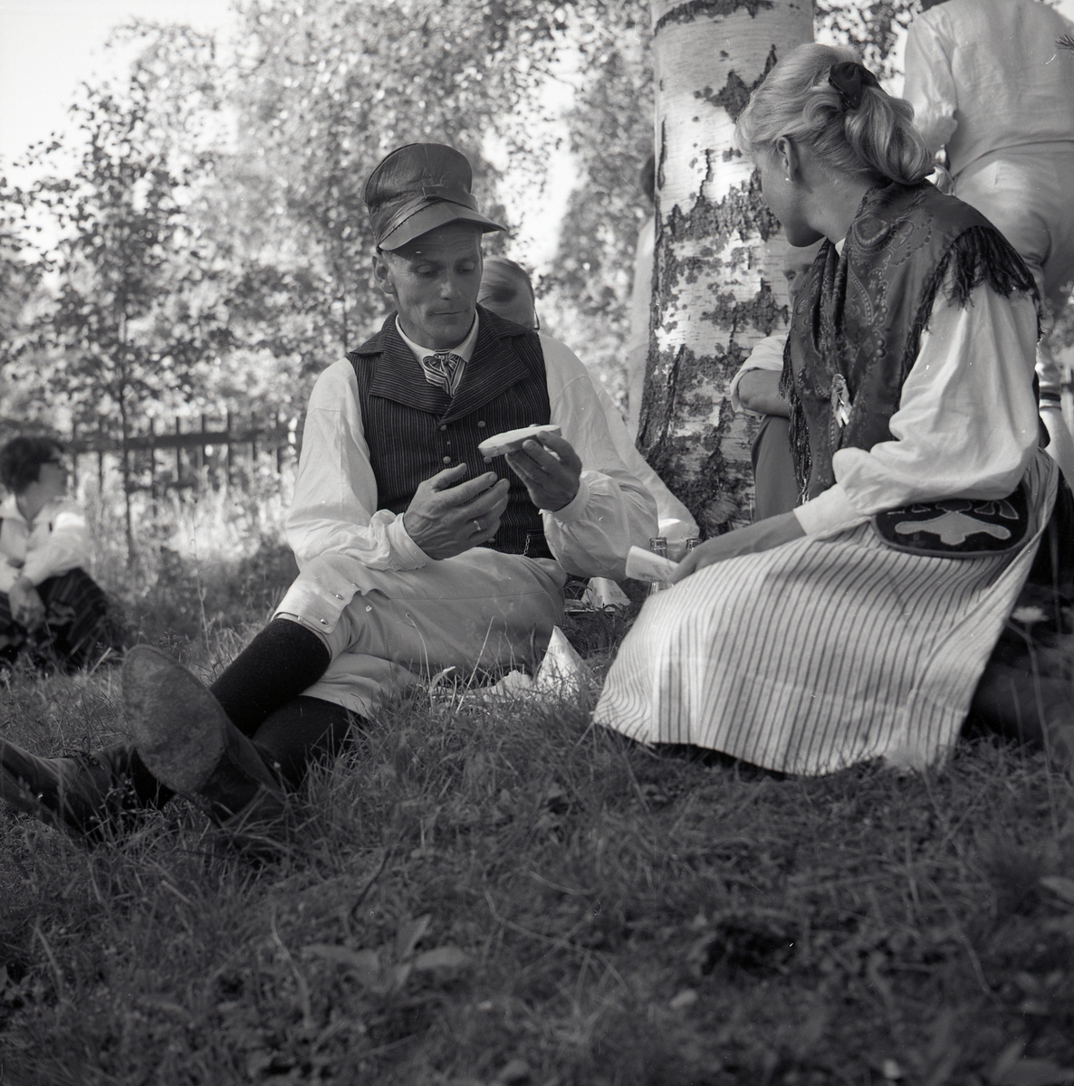 Två dansare sitter och fikar i gräset i skuggan under ett träd vid hälsingehambon, Hårga - Järvsö 1967.