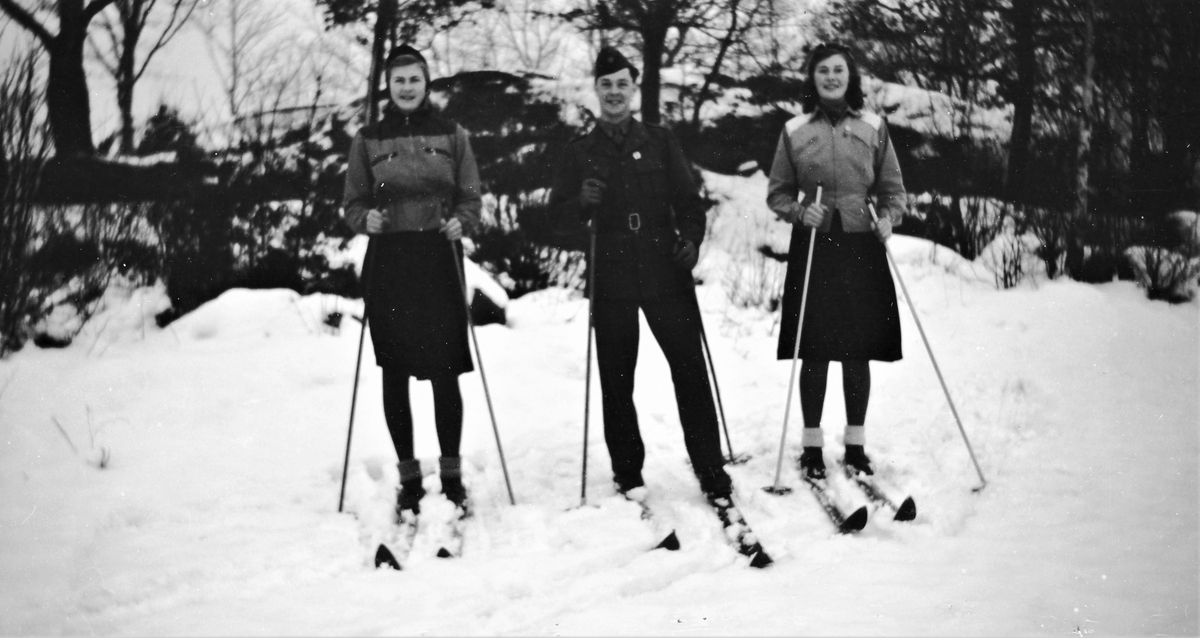 Tre ungdomar på skidor i skogen. Evert Klasson i Albäck står i uniform i mitten med en kvinna på var sida. Tjejerna bär sportiga midjejackor till kjol och mössor, men inga vantar medan Evert har skinnhandskar.