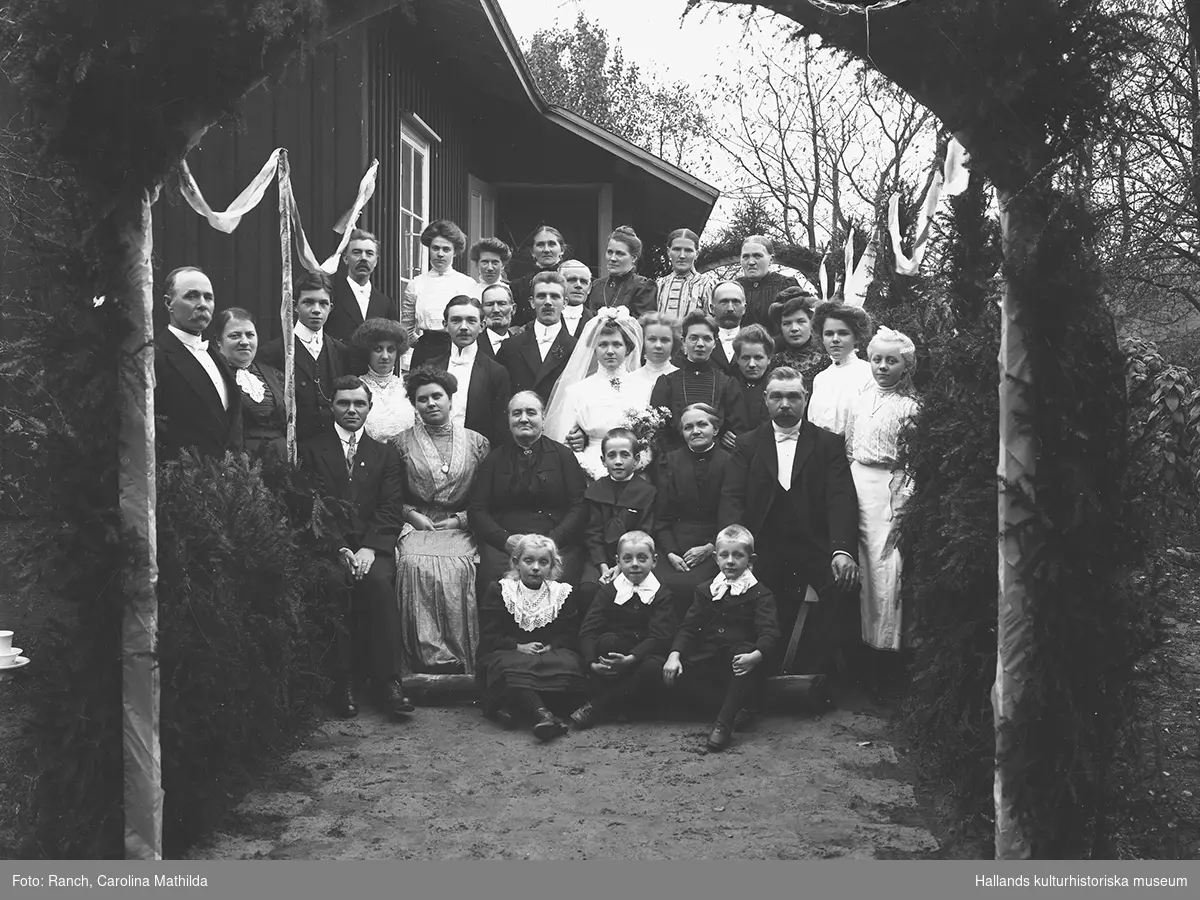 Gruppbild av brudpar med bröllopsgäster. Bilden är tagen utomhus framför ett hus med en äreport i förgrunden. Troligen på landsbygden.
