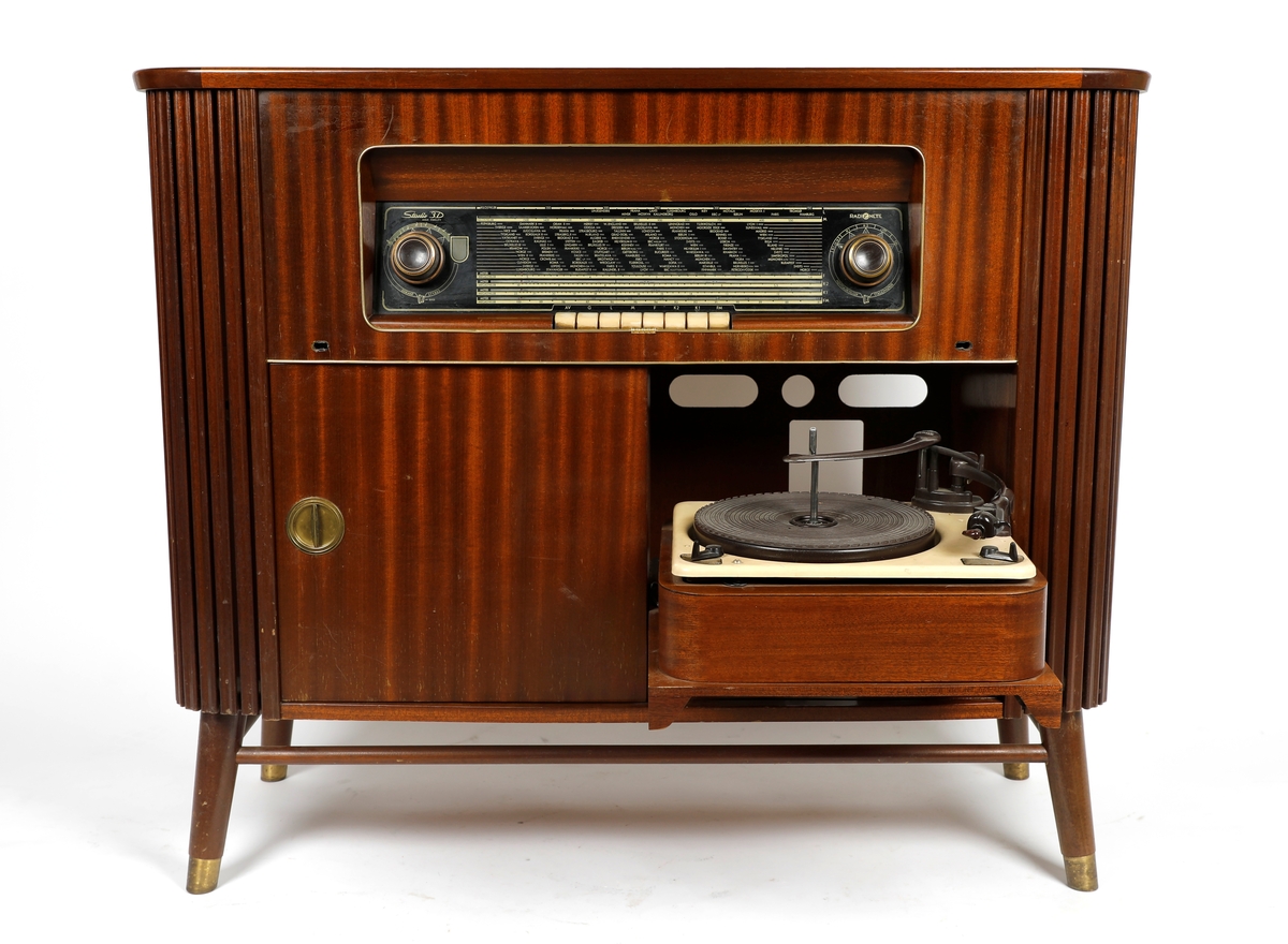 Radiokabinett med en Radinette Studio-3D High Fidelity radio og en Garrard grammofon (platespiller) i høyre side av skapet. To skyvedører i forkant av kabinettet, én nøkkel til skapdør.