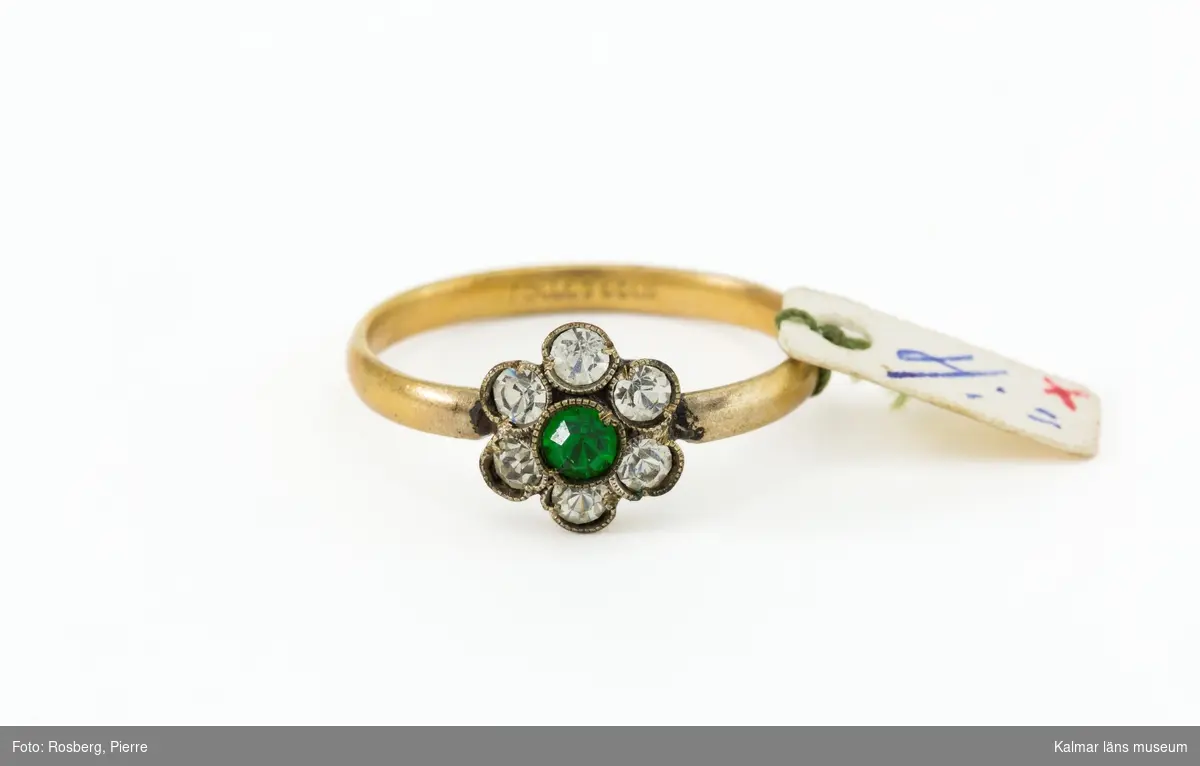 KLM 46368:13:1-2. Ring, fingerring. 2 stycken. Ring av förgylld metall med dekoration i form av en blomma med vita glasstenar med färgad sten i blommans mitt. På ringen en papperslapp: 4 : =. På ring :1 en stämpel: ROLLED GOLD. Ring :2 finns också stämplar men de är så små att de inte går att tyda.