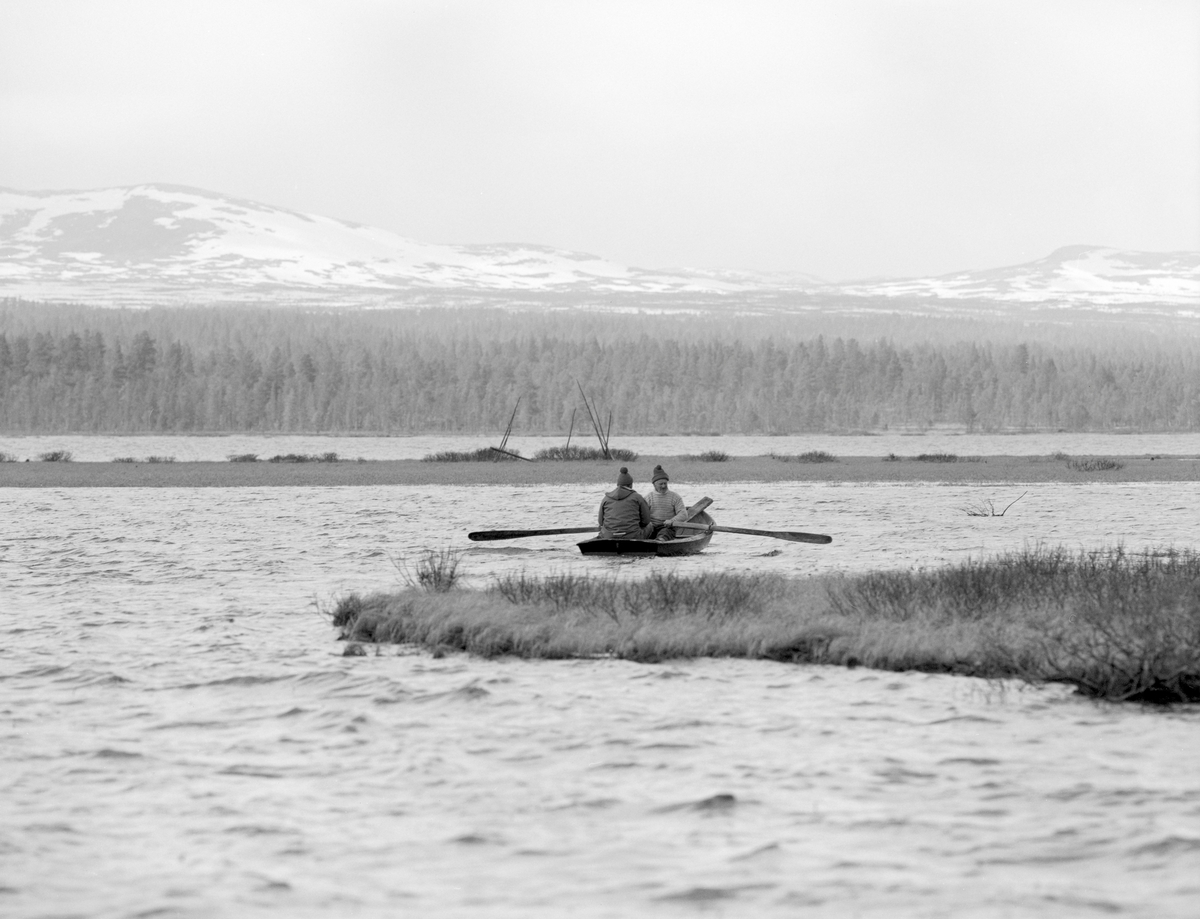 Gjeddefiske i deltaområdet der elva Tufsinga renner ut i innsjøen Femund. Lokaliteten ligger i Tufsingdalen i Os kommune i Nord-Østerdalen, bortimot 700 meter over havet. Fotografiet viser to menn i en klinkbygd trebåt med tverr akterende. Karene skulle trekke gjeddegarn. Vi vet foreløpig ikke hvem mannen i akterenden av båten var. Han som satt ved årene var «gjeddekongen» Joseph Bakken (1898-1989). Gjeddefisket i dette området foregikk i mai måned, like etter at isen på vassdragene hadde smeltet. Da trakk gjedda inn mot de sivbevokste grunnene langs land for å gyte. Der satte fiskerne garn skrått ut fra strandlinja. Det var slike garn de to mennene i båten skulle sette eller trekke da dette fotografiet ble tatt.