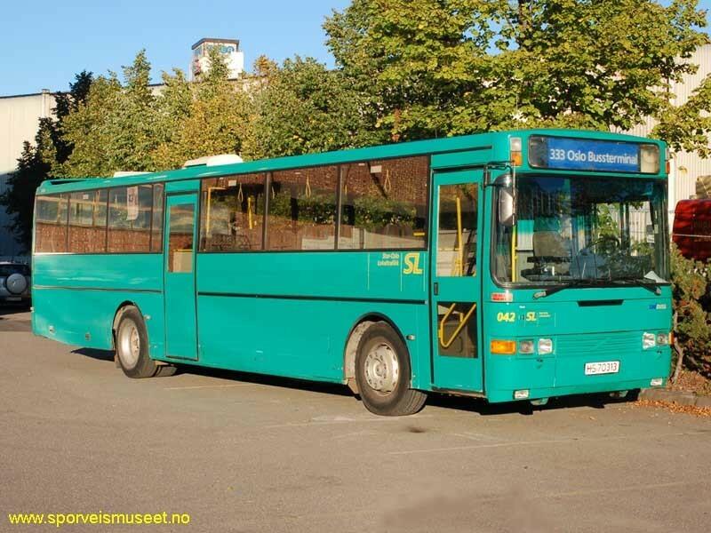 Grønn buss med to enkle inngangsdører, en i front og en i midten. Interiøret består av grå overflater, gule stenger og seter i grått stoff med striper i rød, grønn, svart, gul og blå. 