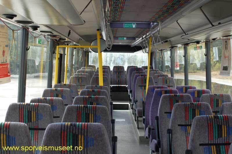 Grønn buss med to enkle inngangsdører, en i front og en i midten. Interiøret består av grå overflater, gule stenger og seter i grått stoff med striper i rød, grønn, svart, gul og blå. 