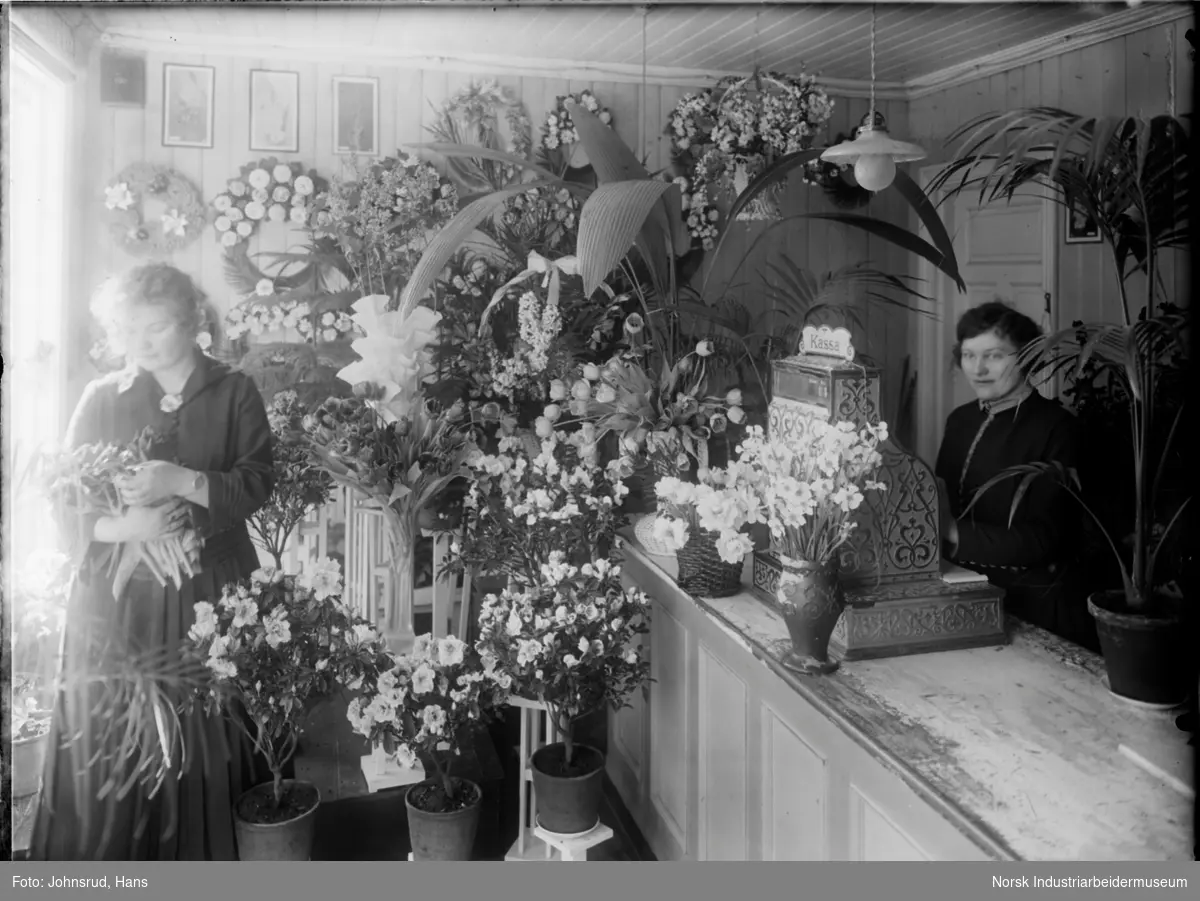 Planter og blomsterbuketter på B. Holter blomsterforretning. Kvinne til venstre med blomster i armene, kvinne til høyre foran en disk med kasseapparat.