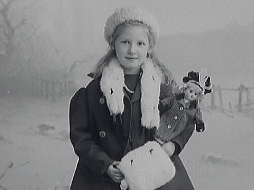 Porträtt av fotografens systerdotter Ingrid Alling med en docka. Hon är vinterklädd med pälsboa och -muff.
