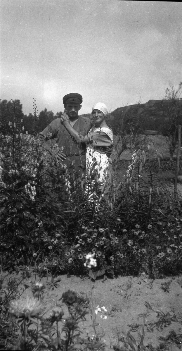 "Kålhage-Karl" med en kvinna vid sin sida bakom en ymnig blomsterrabatt med bland annat stormhattar, liljor och borstnejlikor.