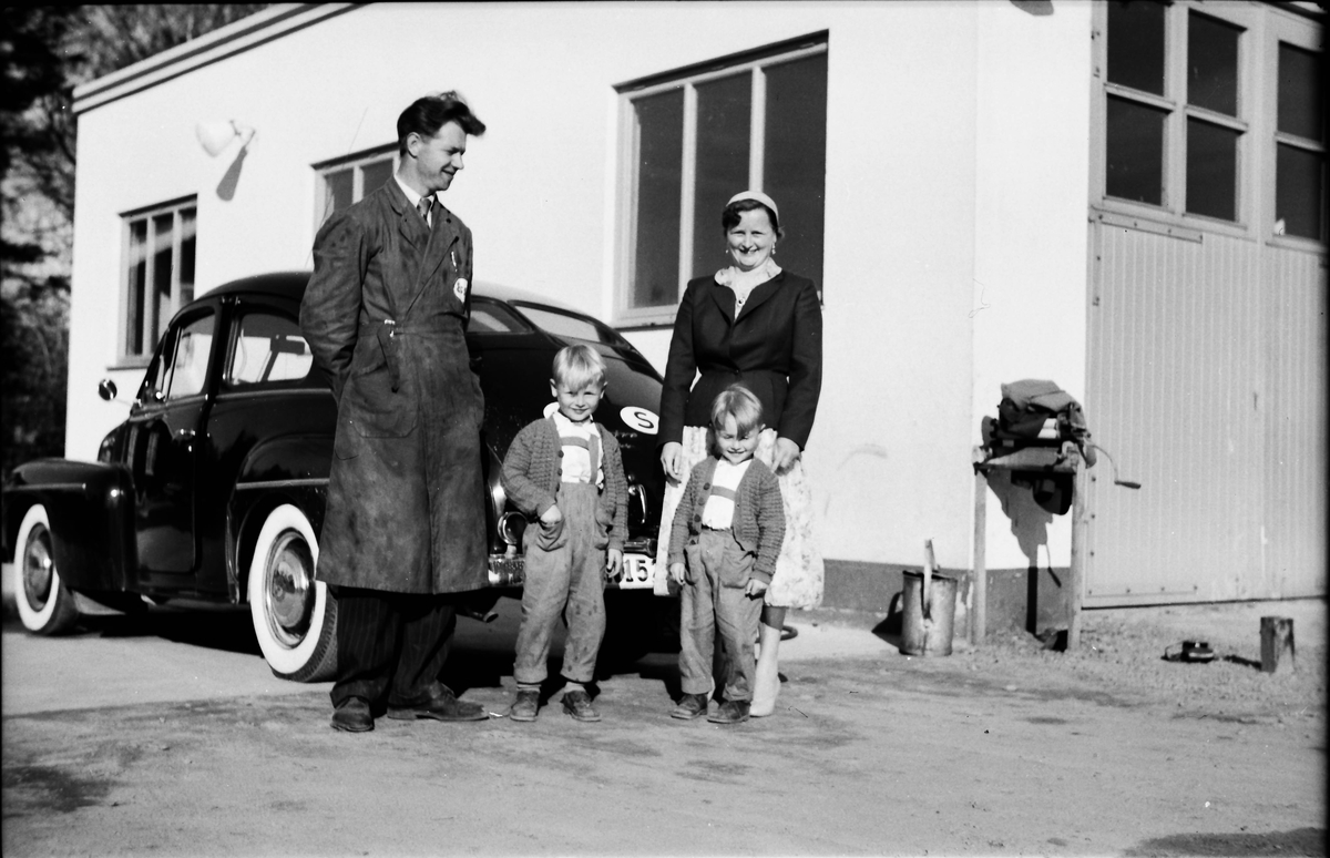 Vid Esso-tappen i Veddige står en skinande Volvo PV med delad bakruta (således äldre än 1955) parkerad. I förgrunden står Henry Svensson i Essorock med frun Dagny och barnen Kennet och Tommy.