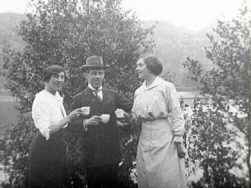 En man omgiven av två kvinnor står med varsin kaffekopp vid en sjö.