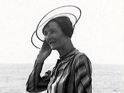En medelålders kvinna klädd i randig klänning och en genomskinlig hatt med två svarta kantcirklar står på stranden vid badplatsen Djupa dräkt i Apelviken. Bild 2 visar närbild på kvinnan och hatten.