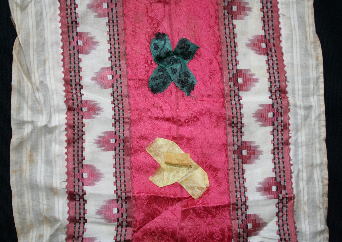 Rektangulär doppåse med sprund bak. Påsens översida av småmönstrad kattun i rosa och vitt, baksida av röd-vitrandigt bomullstyg. Översidan täckt av fyra sammansydda sidenband längs sidorna ikatmönstrade band i vit, rosa, och svart, på mitten två starkrosa blommönstrade band dekorerade med två gula och två mörkgröna sidenbandskryss, i ett av kryssen en knapp med infattad röd glassten. Uptill på tvären ett blekt rosa sidenband. Påsen är på tre sidor kantad med en slät remsa av tunt vitt bomullstyg med satinränder. Nertill rynkat släp av vit tyll broderad i trädning med trianglar och blommor. Foder av vit linnelärft.

/Birgitta Blixt 2021.