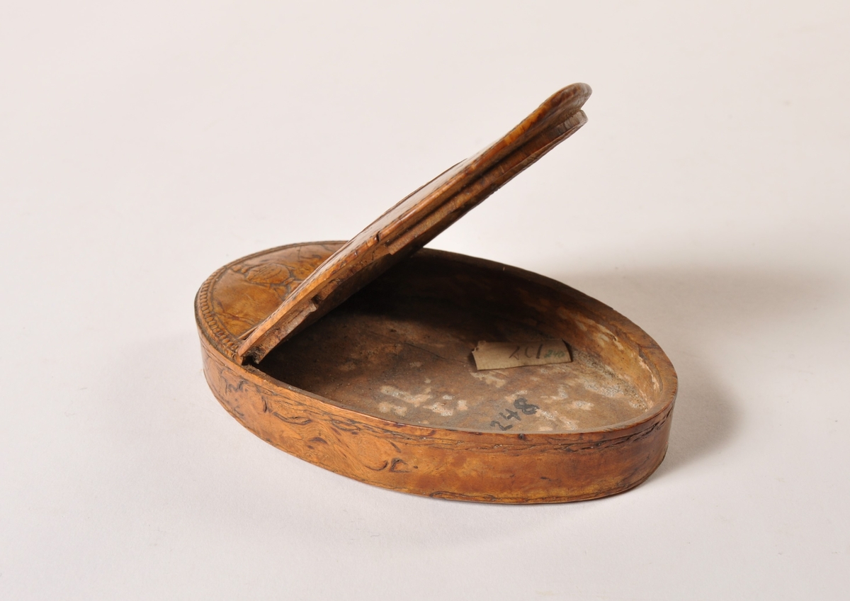 Oval boks av valbjørk, med hengslet lokk. Utskåret mønster på lokk: urne girlander og monogram.