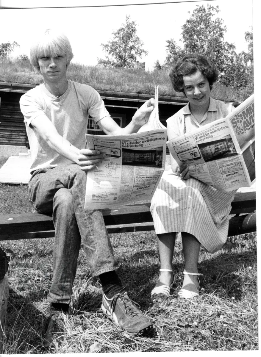 To personer sitter på en benk og leser i avis.