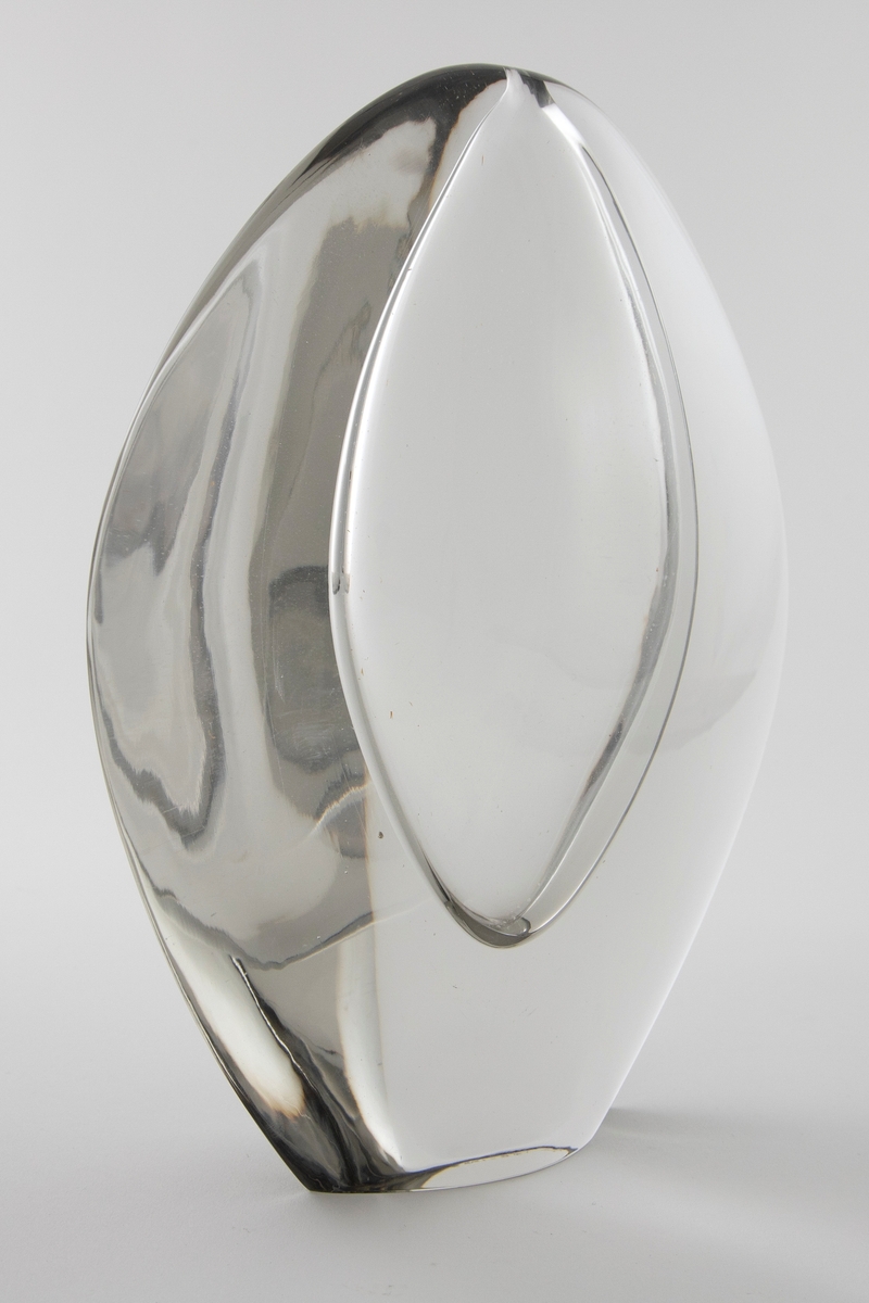 Vase av klart glass med skulpturell utforming. Flattrykket oval hovedform med konvekse sider. Elipseformet hulerom i midten med en liten åpning på toppen.
