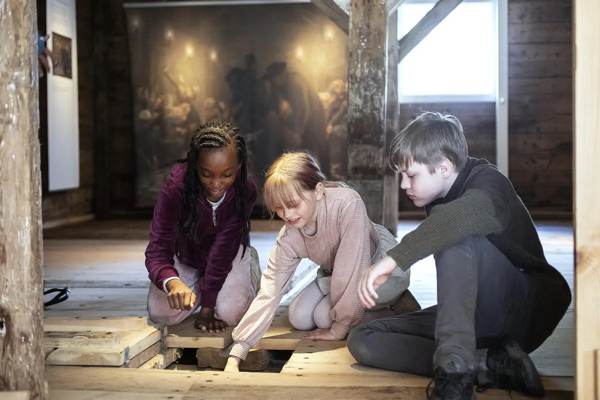 Tre barn sitter på gulvet og studerer luken i gulvet i silderommet på Norges Fiskerimuseum