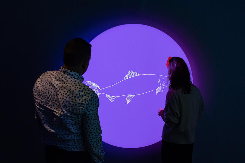 To personer står foran en vegg som viser tegning av en laks inni en lilla sirkel, en del av utstillingen på Storeblå visningssenter for havbruk