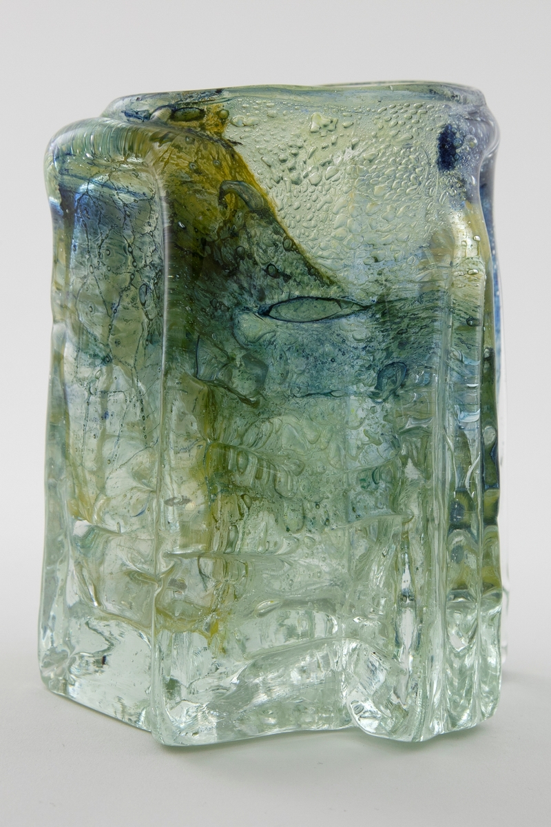 Tilnærmet sylindrisk vase i klart glass med innlagt dekor. Vasens tykke vegger har bølgende former og avsluttes i en sirkulær irregulær munningsrand. Innslag av fargede partier i blå-, gul- og grønnfarget emalje samt luftbobler.