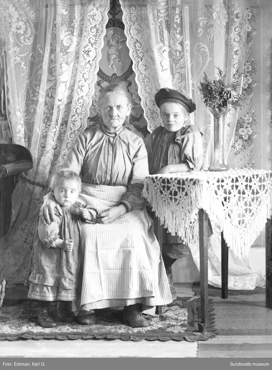 Porträttbild i hemmiljö med en äldre kvinna och två barn, Indal. Ur Karl Eckmans samling.