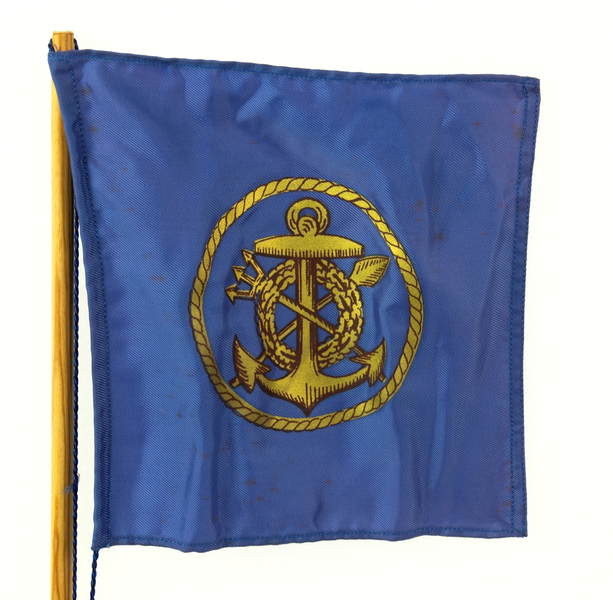 Bordsflagga med Besättningen Rydbergsgastarnas emblem i guld på blå botten. Text skriven på foten: "Ek av regalskeppet Riksäpplet som förliste vid Dalarö Skans år 1676. Stockholm den 31/8 1966. Yngve Lindbäck".