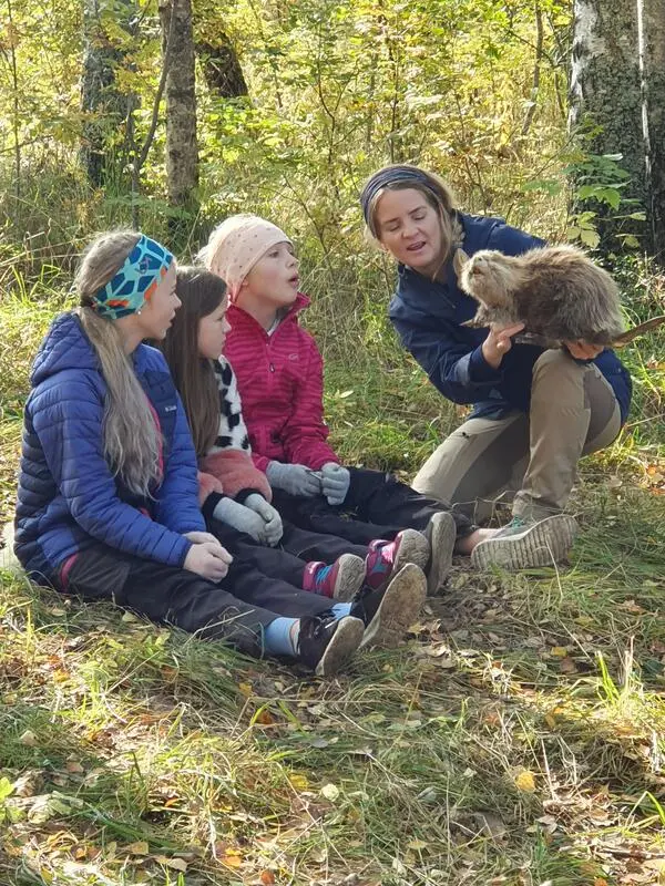 Foto av tre barn som sitter på bakken i skogen sammen med en naturveileder som holder en utstoppa bever