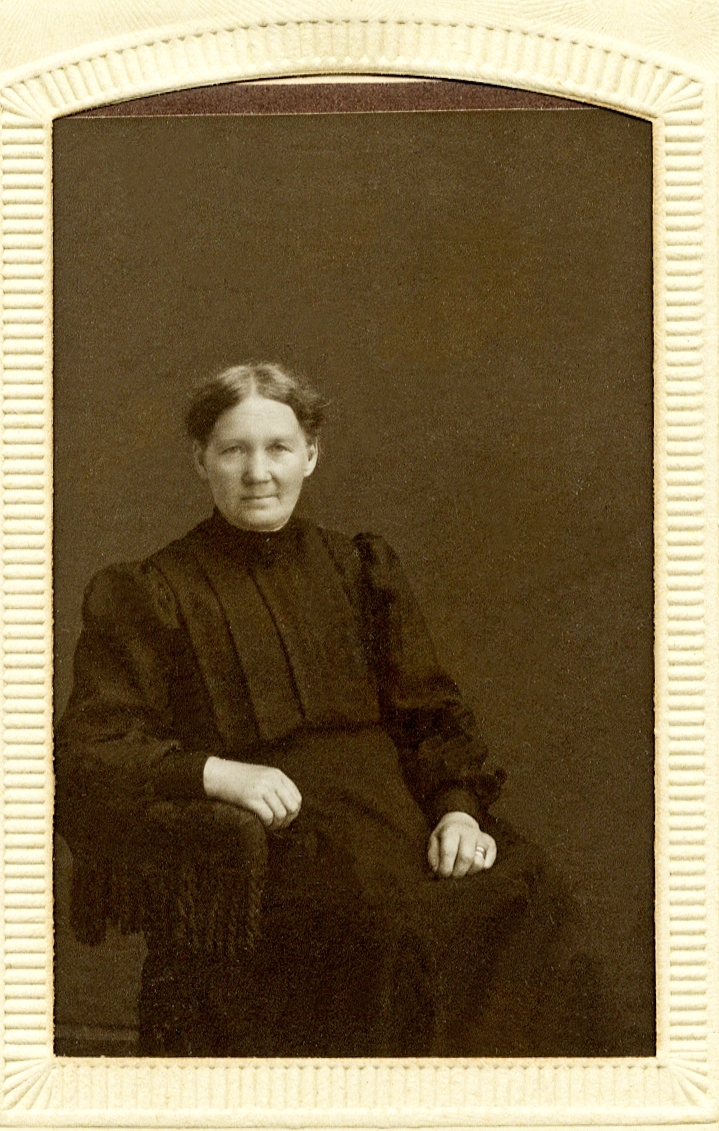 Foto av en sittande äldre kvinna i mörk (svart?) höghalsad klänning. 
Knäbild, halvprofil. Ateljéfoto.