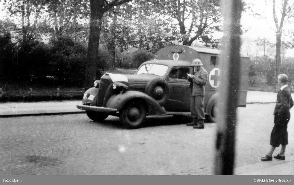 Norsk politisoldat kontrollerer tysk ambulanse, Fredrikstad mai 1945. Bilen er en Chevrolet 1937-modell.  
Antakelig en norsk personbil som har blitt rekvirert av tyskerne og ombygd til nytt formål ved norsk karosserifabrikk. (Ivar Stav 2021)