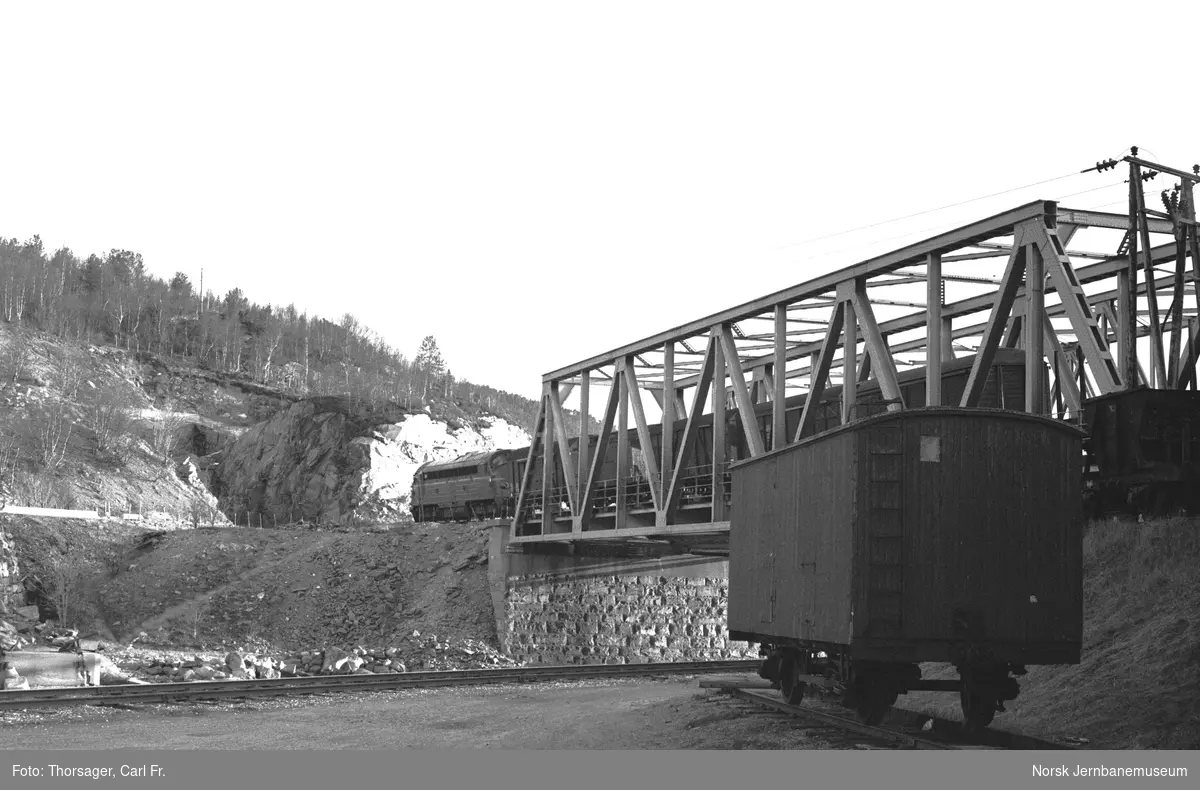 En av Sulitjelmabanens flisvogner litra G nr. 104 eller 103 på Finneid stasjon. I bakgrunnen diesellokomotiv Di 3 med godstog på Finneidstrømmen bru