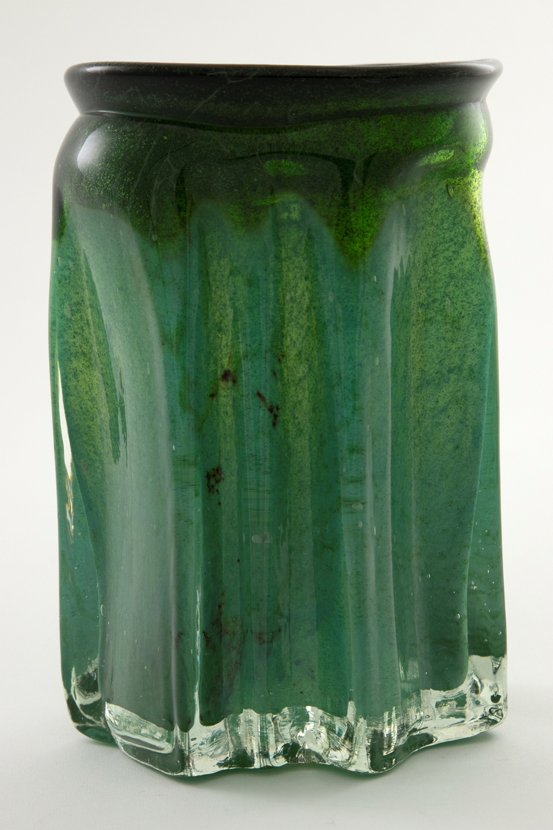 Tilnærmet sylindrisk vase i klart glass med innlagt dekor. Vasens tykke vegger har bølgende former og avsluttes i en sirkulær irregulær munningsrand. Innslag av fargede partier i grønt samt luftbobler og metallspon.