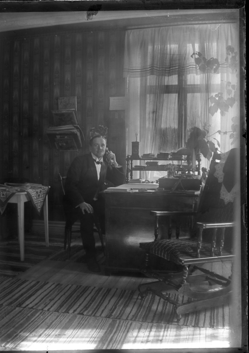 En man sitter vid ett skrivbord och talar i telefon. I förgrunden står en gungstol till höger. Solen skiner in från samma håll.