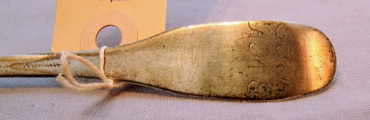 Matsked i silver tillverkad av C.G. Wetterström år 1756. Inristade bokstäver på två rader bak på skaftet. Den översta radden är svårtydd men den nedre lyder "A M E". Skeden är odekorerad.