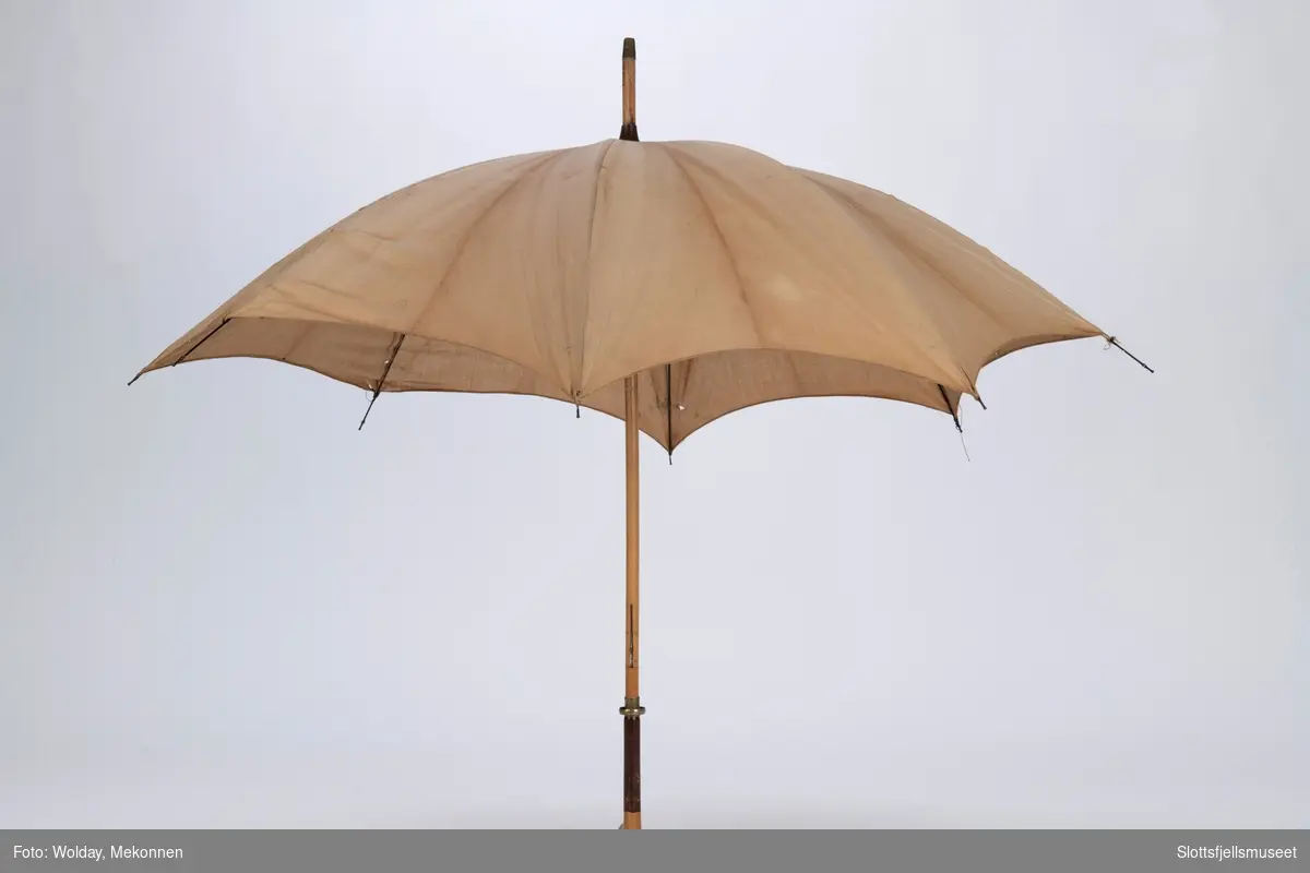 Paraply med 8 spiler i metall og treskaft. Paraply er trukket med lyst bomullsstoff. Treskaftet ender i en kølleformet håndtak. 
