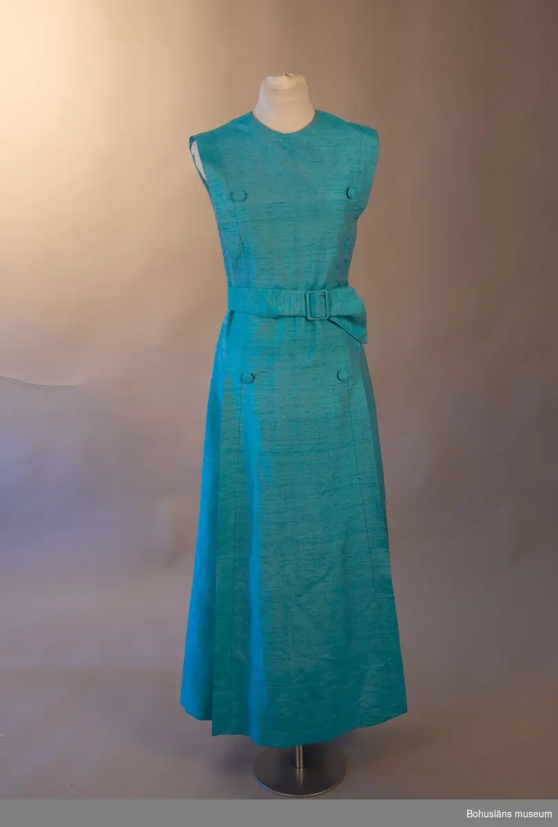 Turkos sidenklänning med ett hängande del framtill fästad i klänningens axel och halsdel. Tyget hålls in med skärpet som knytes i midjan. Skärpet bidrar till klänningens fall och form.