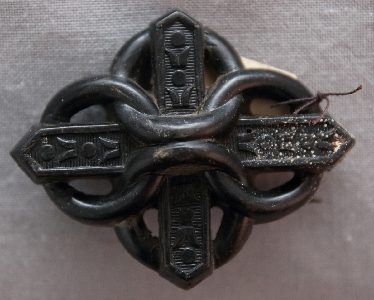 Svart brosch i ebonit i form av ett likarmat kors omflätat av fyra ringar.