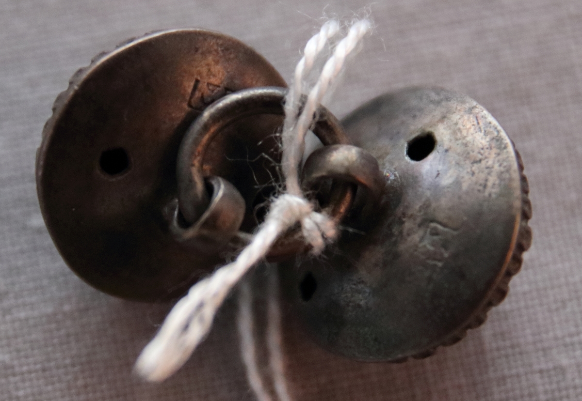 Två buckleformade knappar av tenn, vars märlor sammanlänkats av en mindre ring. Dekor i relief föreställande blommor i mitten av knapparna och en bård längs kanterna.